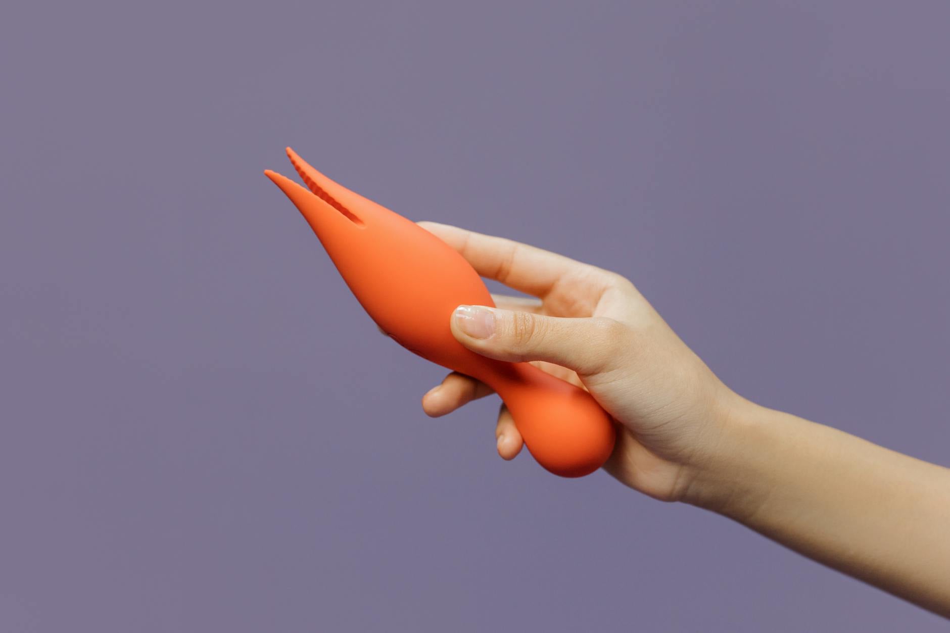 Een close-up van een hand met een oranje vibrator tegen een minimalistische paarse achtergrond, toont moderne sensualiteit